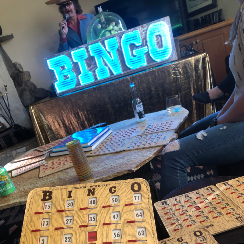 A bingo card being held in front of a neon sign that reads, "BINGO"