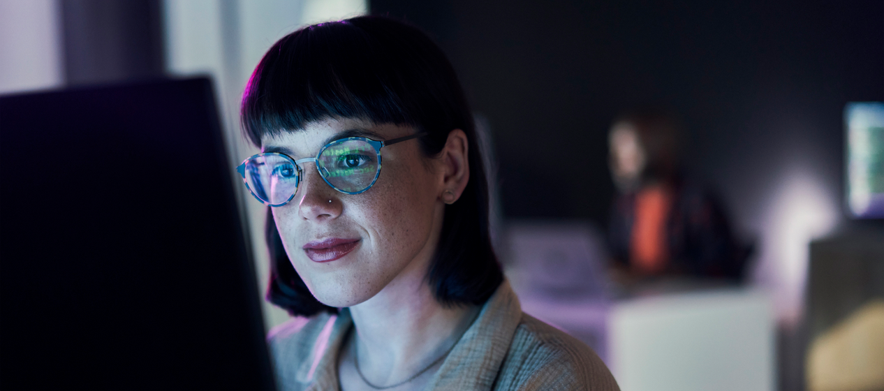A woman with glasses smiles slightly while working at a computer in a blue-lit modern office.