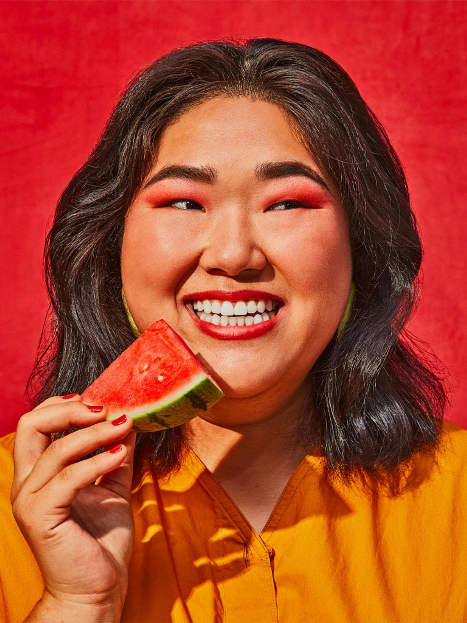 A joyful woman biting a watermelon slice against a red backdrop.