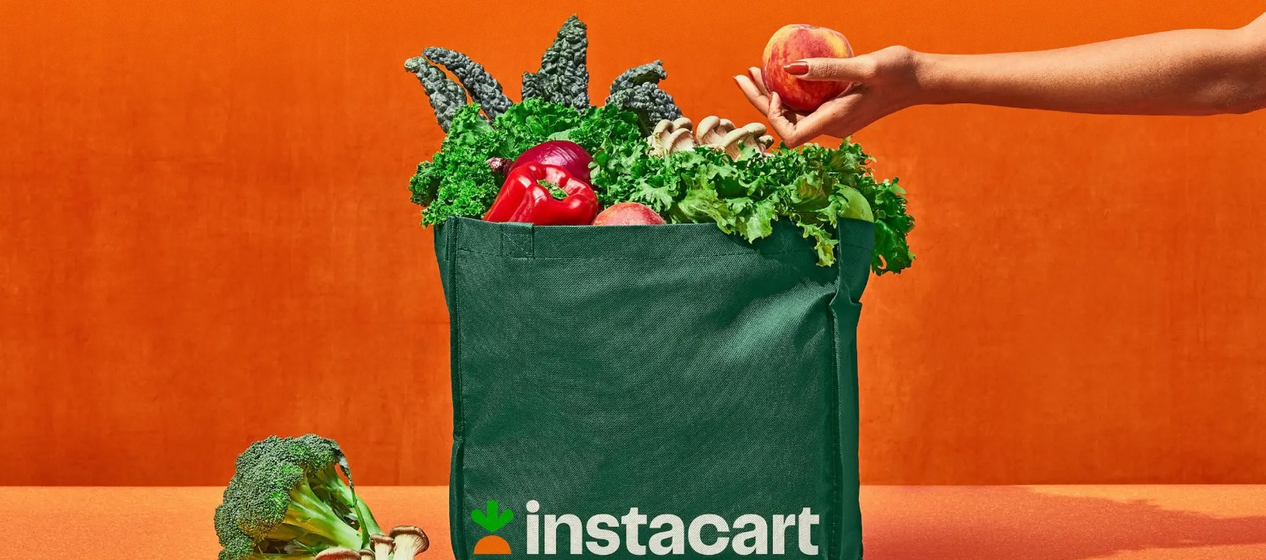 A hand adds an apple to a green Instacart bag full of fresh produce, with an orange backdrop.