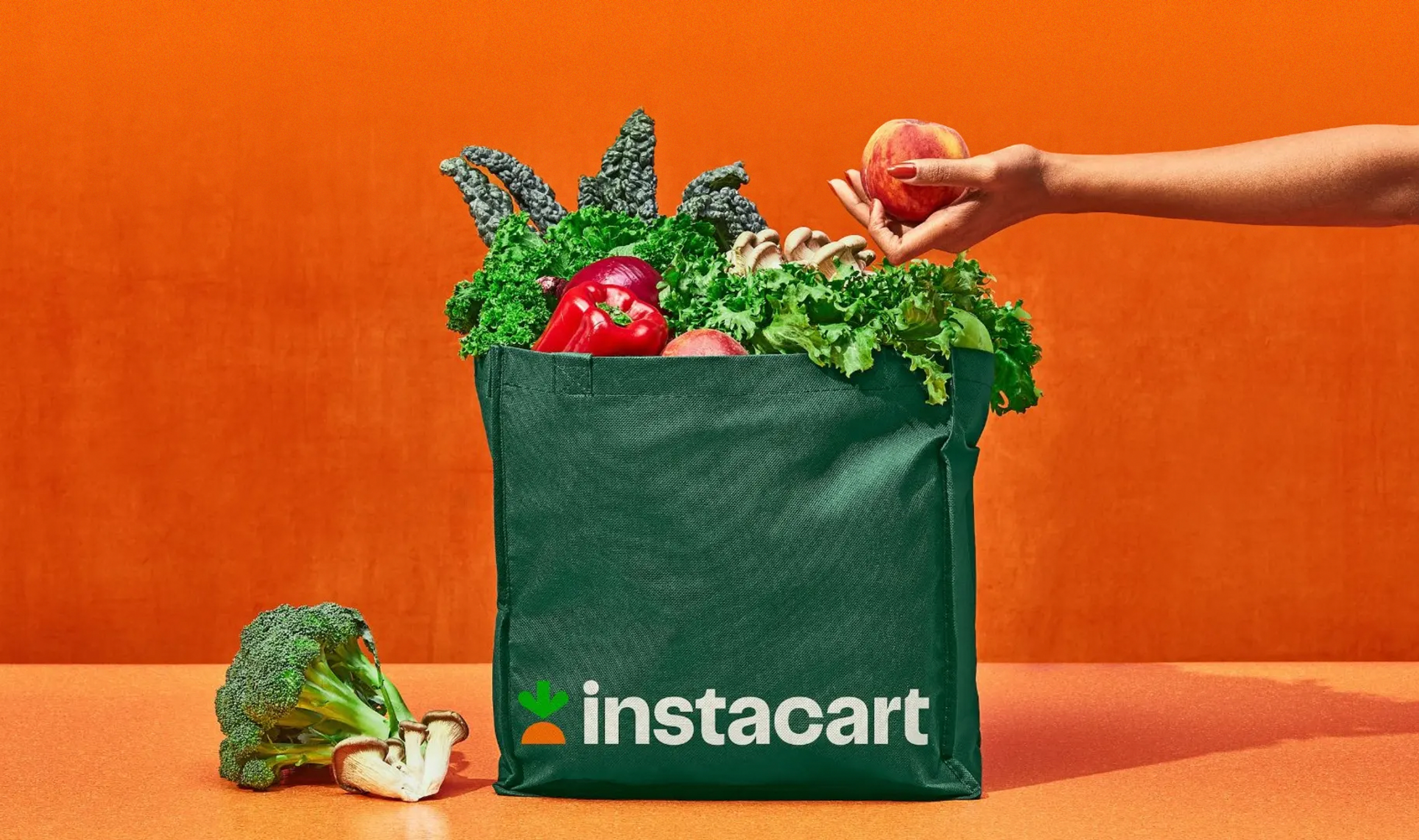 A hand adds an apple to a green Instacart bag full of fresh produce, with an orange backdrop.